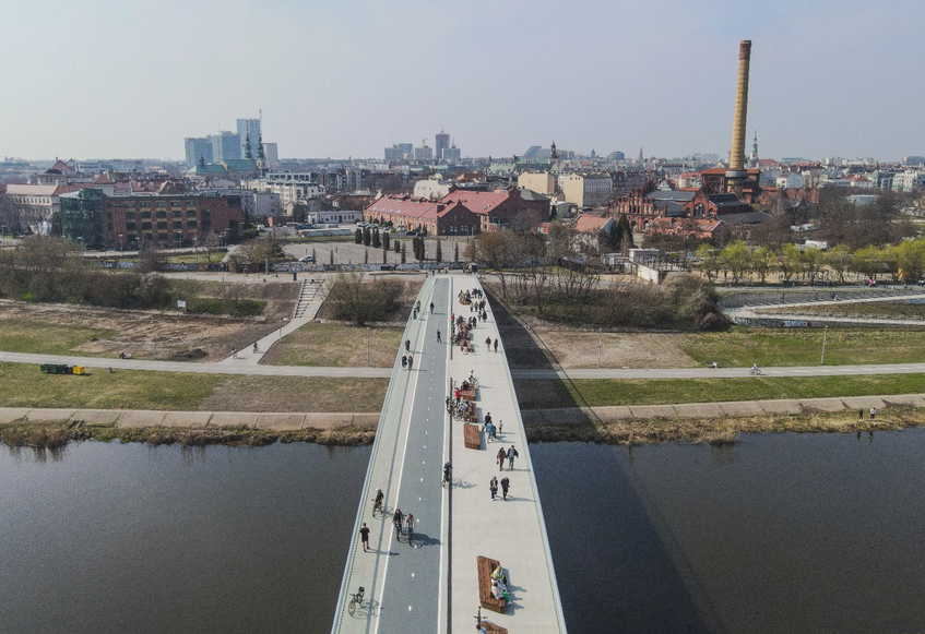 Fot. Miasto Poznań wiodk z lotu ptaka na kładkę pieszo-rowerową nad rzeką za nią na brzegu widoczne zabudowania przemysłowe jeszcz dalek wieżowce