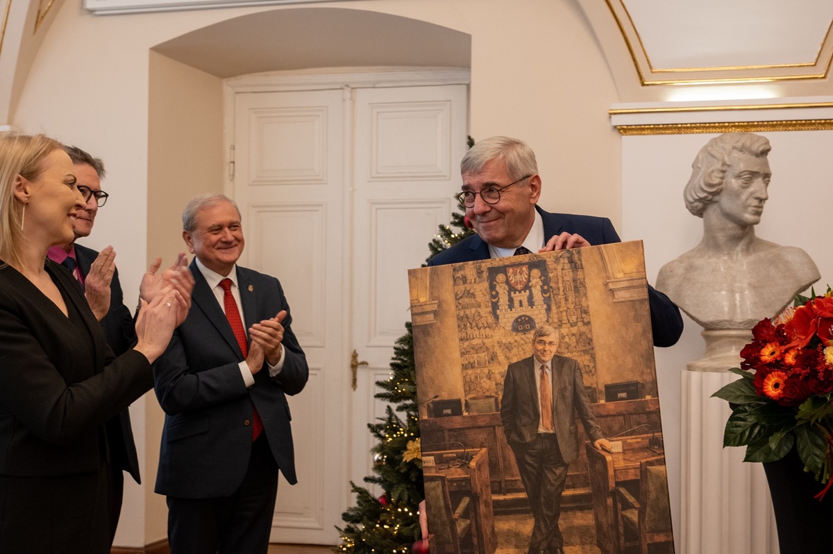 Mężczyzna w średnim wieku ubrany w garnitur, w okularach trzyma w rękach swój portret, obok inne osoby składają mu życzenia