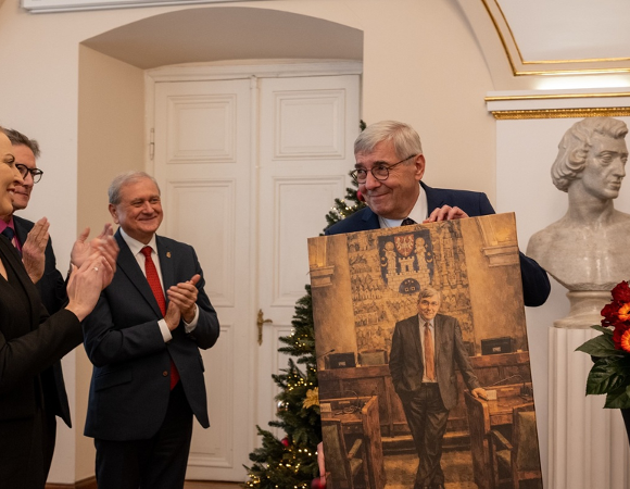 Mężczyzna w średnim wieku ubrany w garnitur, w okularach trzyma w rękach swój portret, obok inne osoby składają mu życzenia