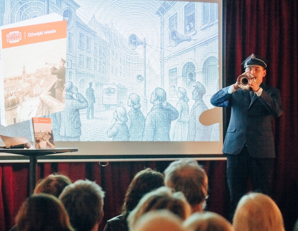 na zdjęciu mężczyna w mundurze grający na trąbce przed widownią, za nim na dużym ekranie wyświetlana jest prezentacja multimedialna - więcej w treści artykułu