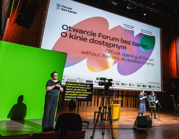 Na zdjęciu tłumacz polskiego języka migowego na zielonym tle. W tle plansza z informacją o otwarciu forum.