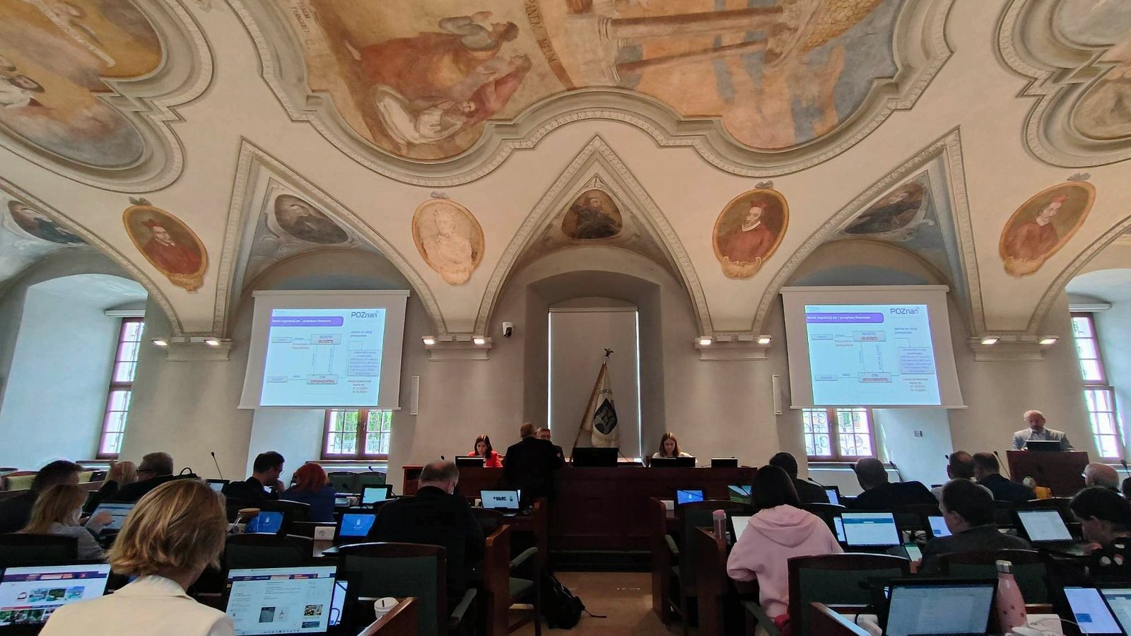 Radni poznańscy w czasie sesji, w głębi stół prezydialny. Nad radnymi sklepienie z odsłoniętymi freskami