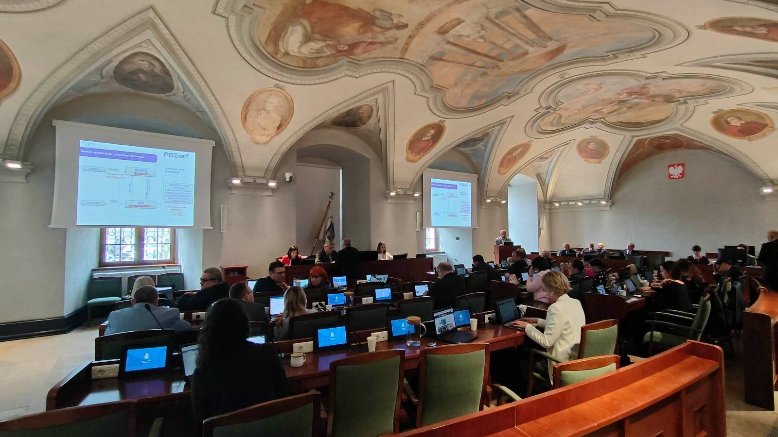 Radni poznańscy w czasie sesji, w głębi stół prezydialny. Nad radnymi sklepienie z odsłoniętymi freskami