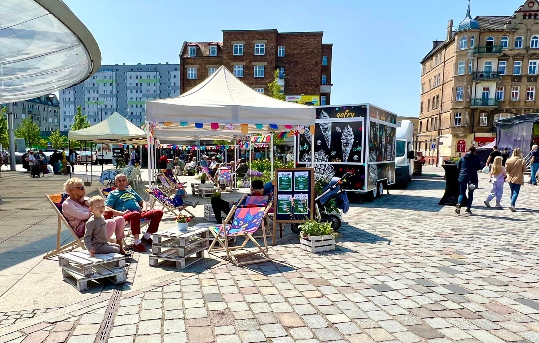 Słonieczny dzień na miejskim rynku, ludzie siedzą na leżakach, za nimi stoiska gastronomiczne w przyczepach tzw. food trucki - grafika artykułu