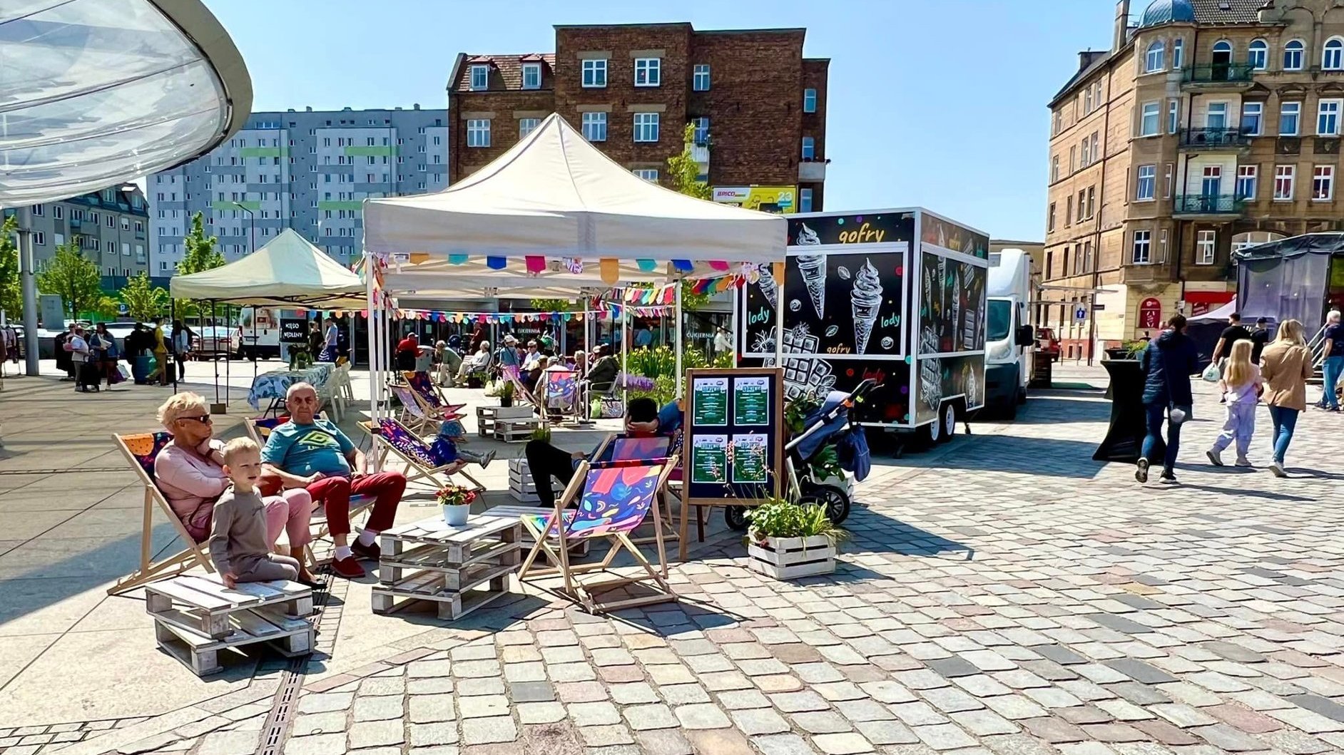 Słonieczny dzień na miejskim rynku, ludzie siedzą na leżakach, za nimi stoiska gastronomiczne w przyczepach tzw. food trucki