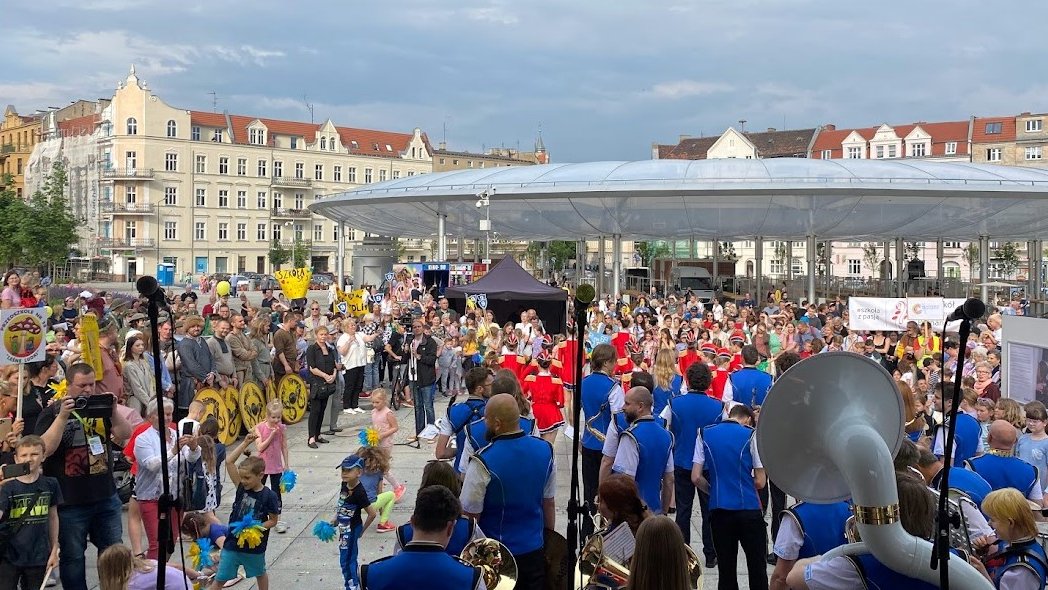 Orkiestra dęta w niebieskich kamizelkach występuje przed liczną grupą mieszkańców na miejskim rynku