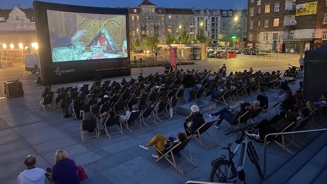 Wieczór na miejskim rynku, ludzie siedzą na krzesełkach i oglądają film na ekranie wielkoformatowym