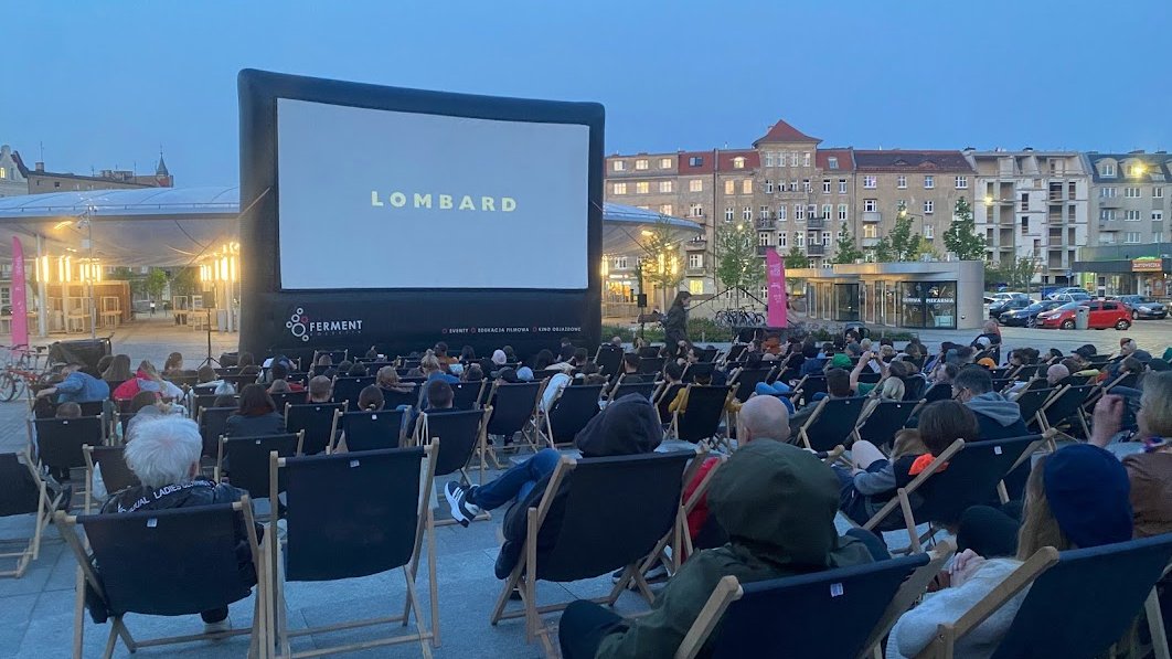 Wieczór na miejskim rynku, ludzie siedzą na krzesełkach i czekają na pokaz filmu Lombard na ekranie wielkoformatowym