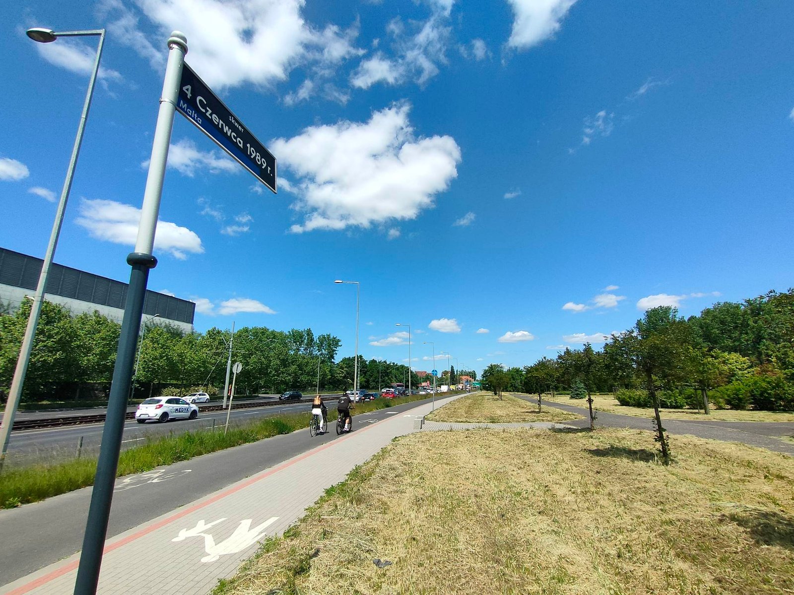 Ścieżka rowerowa przy ruchliwej ulicy, po lewej tabliczka z nazwą skweru, po prawej rozległy i zadbany tereni zielony z drzewami i krzewami - grafika artykułu