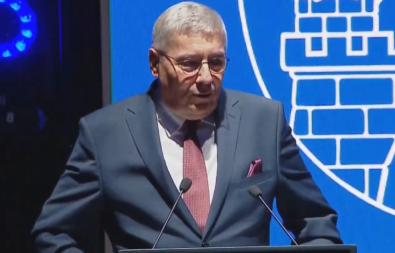 Mężczyzna w garniturze i okularach przy mównicy, za nim fragment herbu Miasta Poznania - grafika artykułu