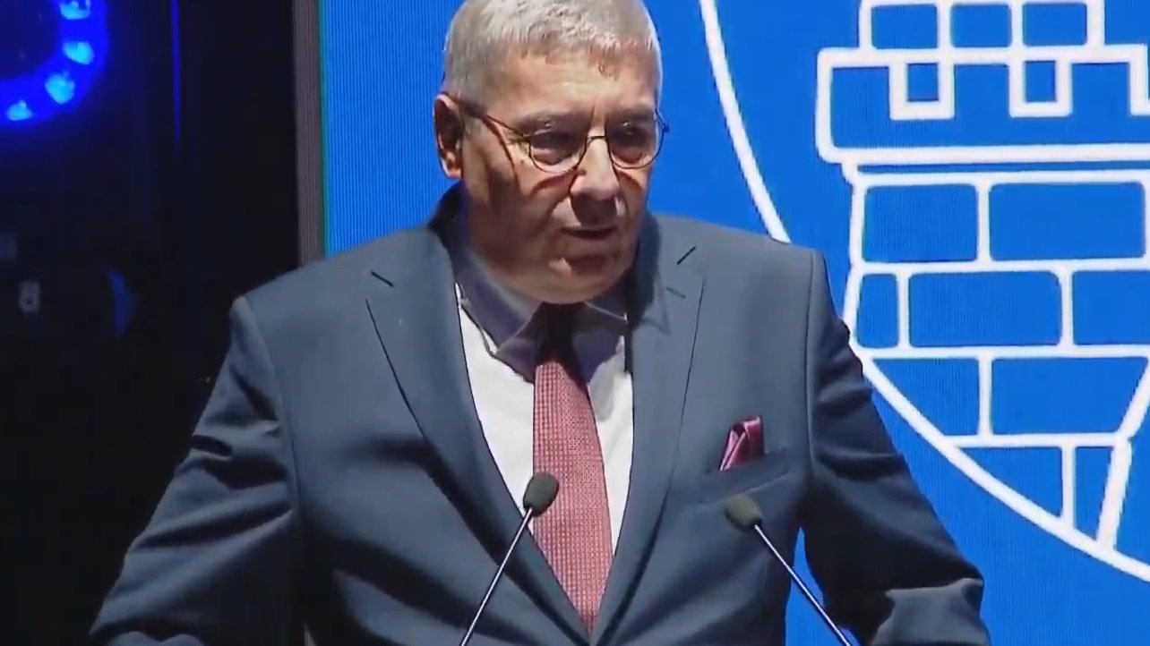 Mężczyzna w garniturze i okularach przy mównicy, za nim fragment herbu Miasta Poznania