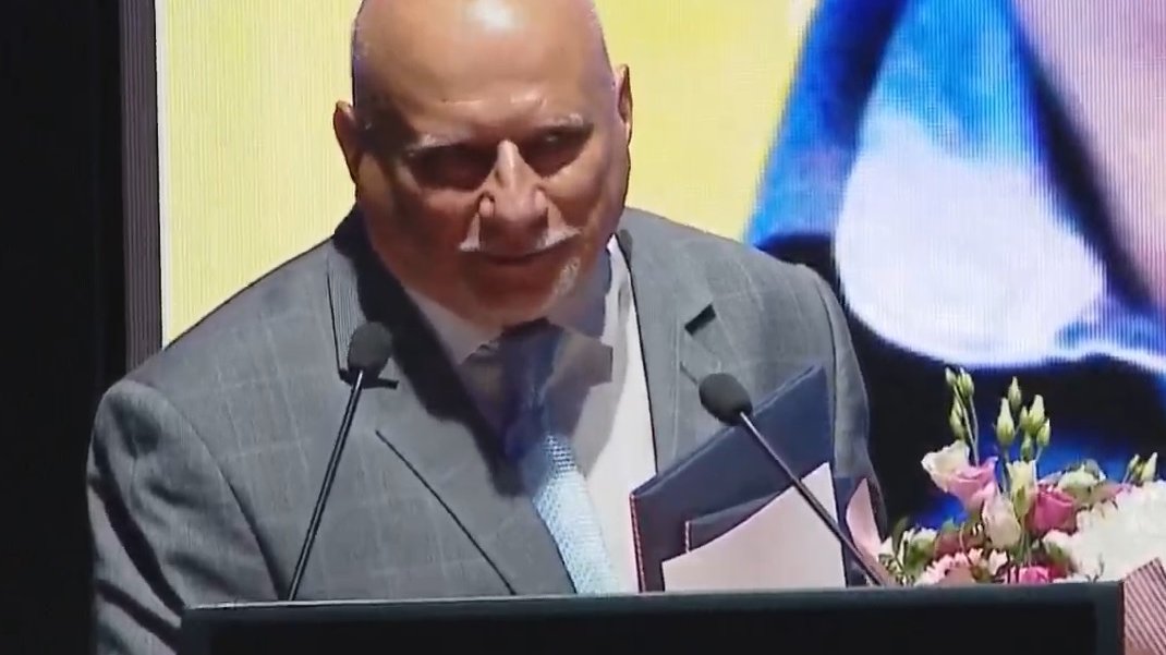 Mężcyzzna w garniturze, z dyplomem i bukietem kwiatów w ręce, przy mównicy, mówi do mikrofonu, dziekując za przyznane wyróżnienie