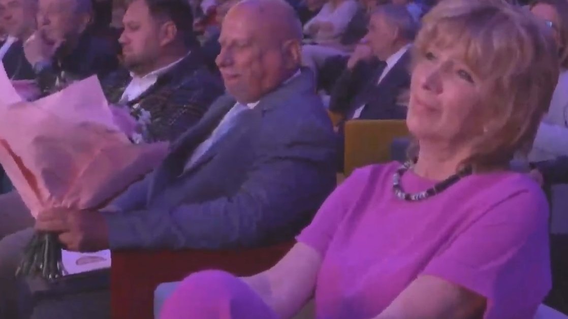 Uśmiechnięta kobieta w różowym kombinezonie-garniturze siedzi na fotelu w sali pełnej gości