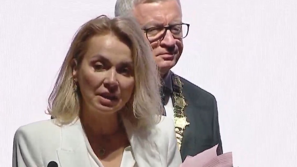 Kobieta w białej marynarce, z kawiatami, przy mównicy. Za nią prezydent miasta z łańcuchem na piersiach