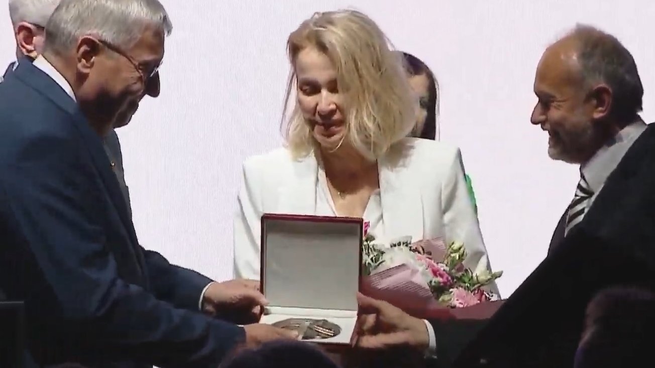 Kobieta w białej marynarce odbiera medal od mężczyzny w szarym garniturze na uroczystej sesji