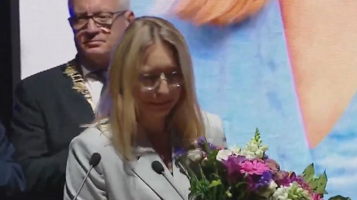 Kobieta w okularach, z kwiatami w ręku, przy mikrofonie. Za nią prezydent miasta z łańcuchem na piersiach