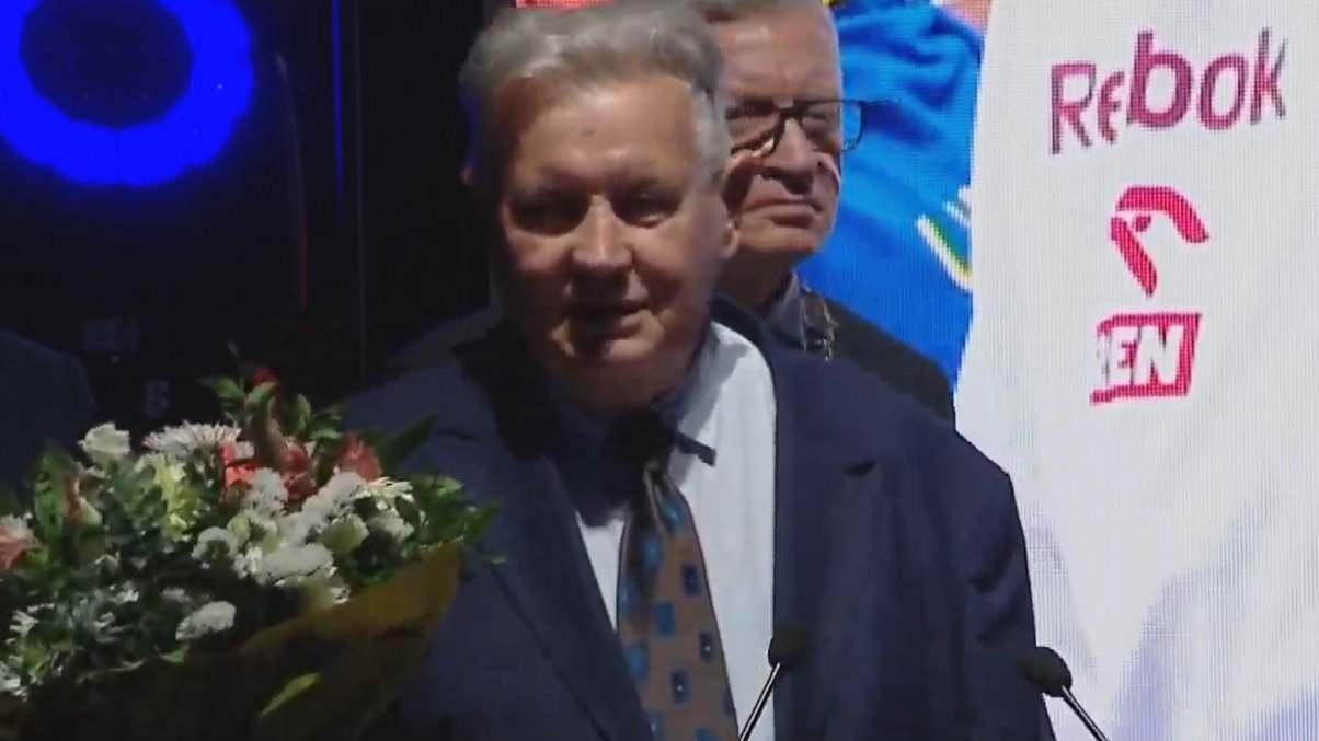 Uśmiechnięty mężczyzna w marynarce, z kwiatami w ręku, przy mikrofonie. Za nim prezydent miasta