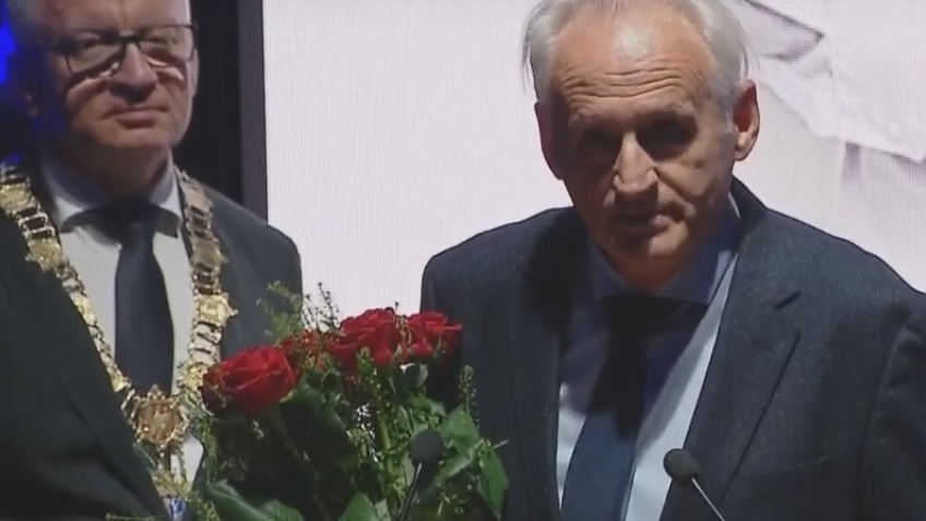 Przy mównicy mężczyzna w garniturze z kwiatami w rękach, za nim prezydent miasta z łańcuchem na piersiach