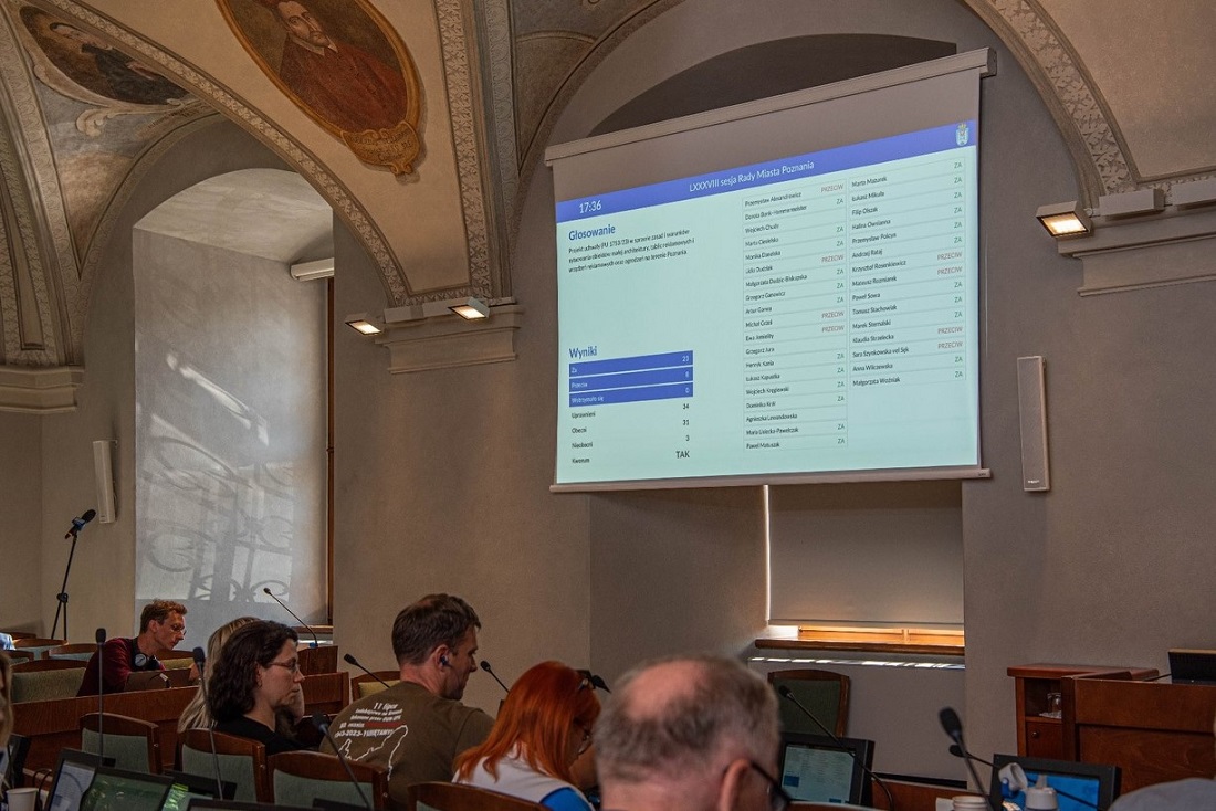 grupa osób kobiety i mężczyźni, siedzą przy pulpitach z ekranami i mikrofonami, w sali z jasnymi ścinami i sufutem zdobionym freskami - grafika artykułu