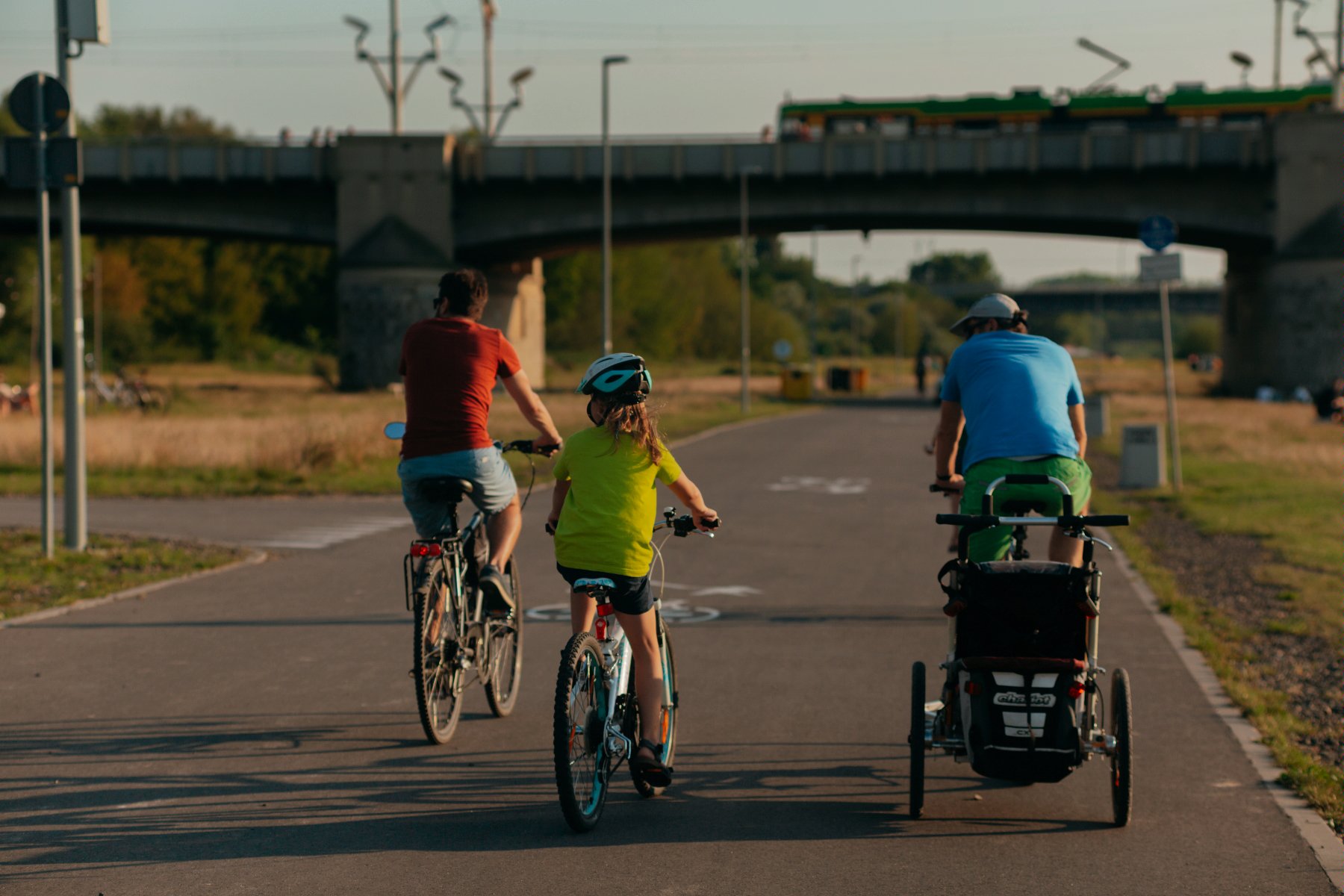 Rodzinny przejazd rowerem po trasie asfaltowej w świetle zachodzącego słońca, mężczyzna na rowerze z przyczepką dla dziecko, starsze dziecko i jeszcze jeden rowerzysta na swoich rowerach - grafika artykułu