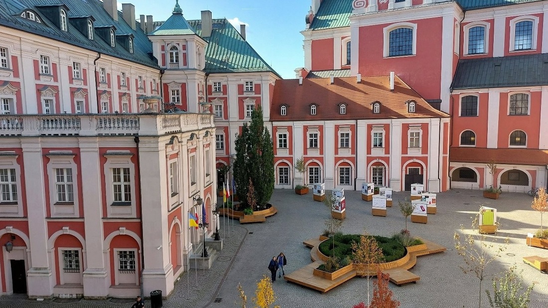 dziedziniec z dużym kwietnikiem na środku, wokół zespół budynków w kolorach czerwonych i białych - grafika artykułu