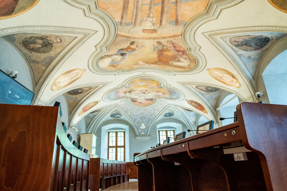 Sklepienie sali ozdobione barwnymi freskami, w dole stanowiska dla radnych miejskich wyposażone w monitory i mikrofony - grafika artykułu