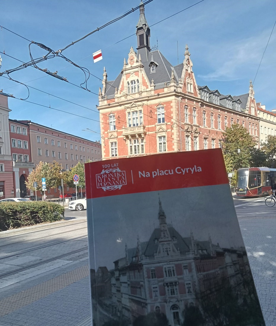Książka na tle placu Cyryla Ratajskiego tytuł "Na placu Cyryla" za książką widać ten sam budynek co na okładce na ulicy widać samochód, tramwaj i rower - grafika artykułu