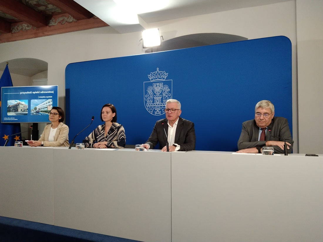 Za stołem konferencyjnym siedzą dwie kobiety i dwóch mężczyzn za nimi niebeiskie tło z herbem Poznania - grafika artykułu