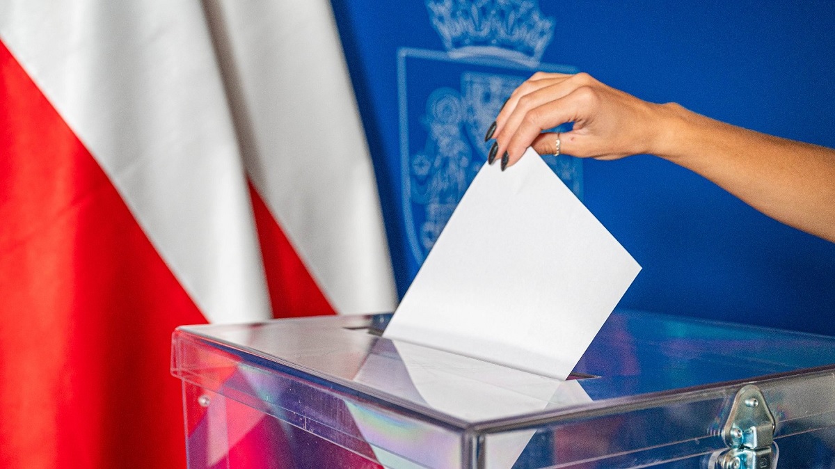 Ręka wrzucająca kartkę papieru do urny wyborczej. W tle flagi Polski i herb Poznania. - grafika artykułu