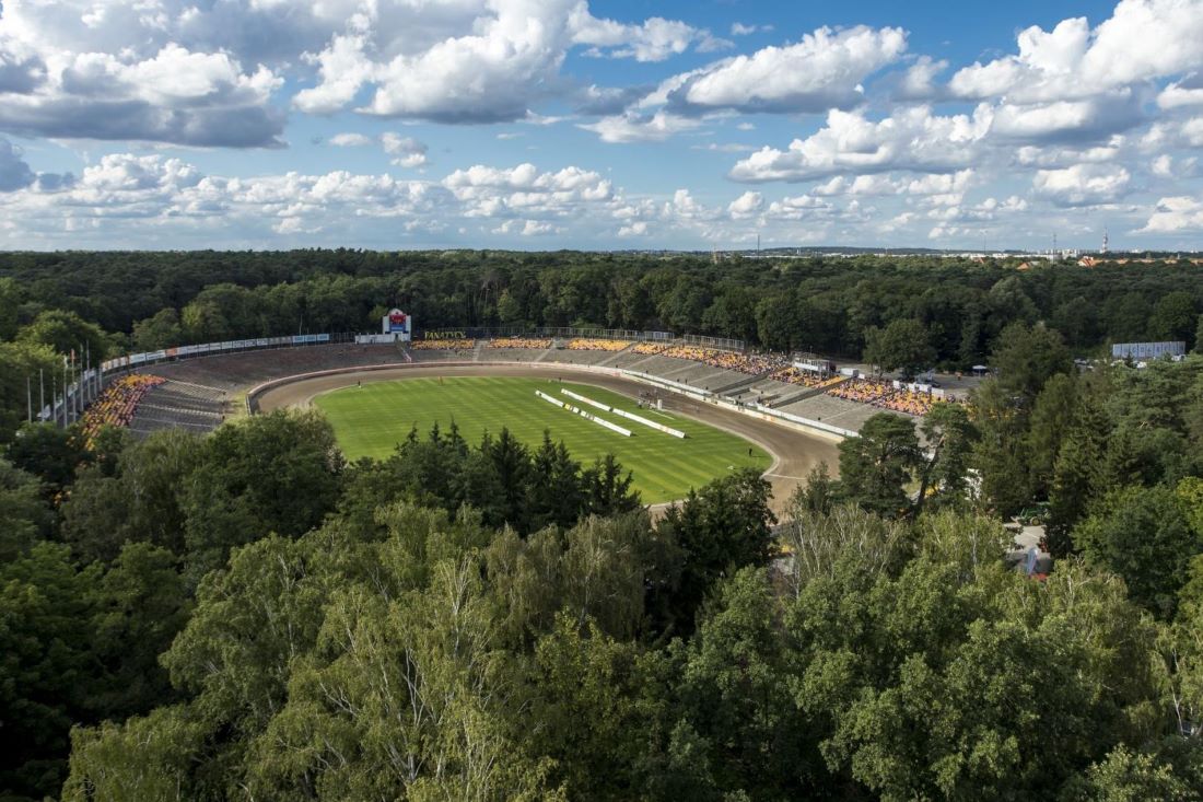 widok z wysokości, stadion położony wśród drzew, widoczne trybuny otaczające murawę oraz tor żużlowy - grafika artykułu