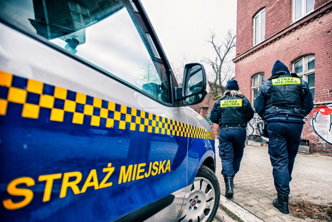 fragment samochodu straży miejskiej, obok dwoje strażników miejskich, kobieta i mężczyzna w umundurowaniu, widoczni tyłem, w tle kamienica z czerwonej cegły - grafika artykułu