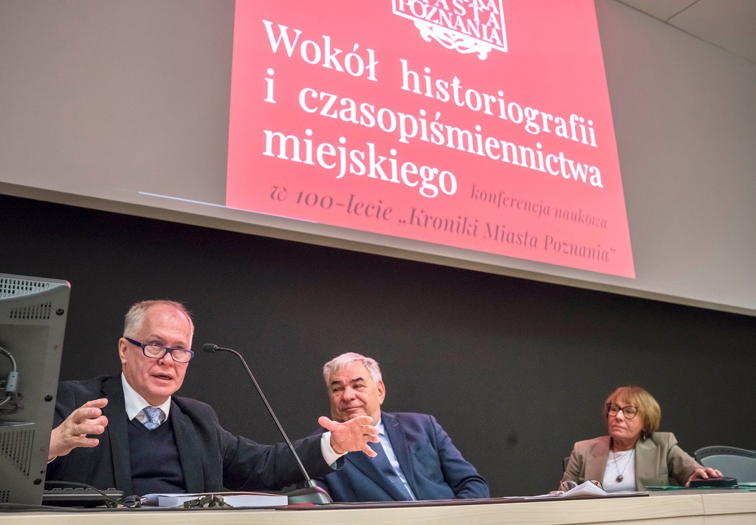 Za stołem konferencyjnym siedzą trzy osoby, dwóch mężczyzn i kobieta, mężczyzna z lewej strony ma przed sobą mikrofon, przemawia, ma rozłożone ręce, okulary, pozostałe osoby patrzą w jego stronę, nad nimi ekran z wyświetlonym tytułem konferencji "Wokół historiografii miast i czasopiśmiennictwa miejskiego" konferencja naukowa w 100-lecie Kroniki Miasta Poznania - grafika artykułu