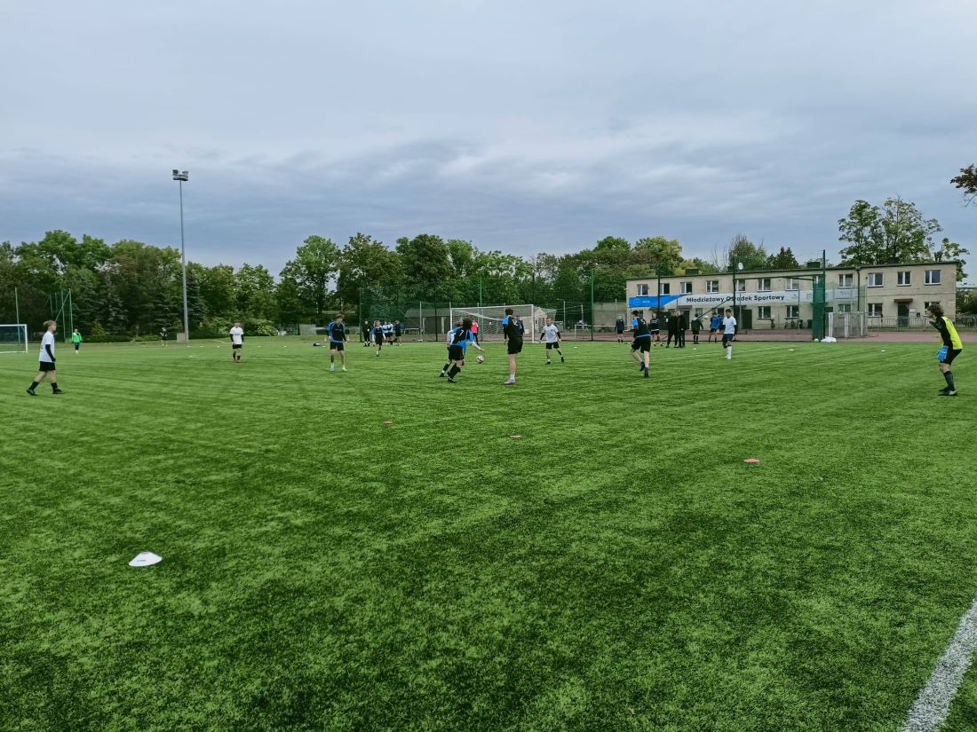 boisko sportowe, zawodnicy grają w piłkę, za boiskiem drzewa i dwukondygnacyjny budynek - grafika artykułu