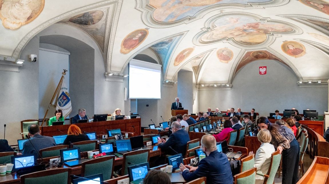Sala sesyjna Rady Miasta Poznania, trwa sesja, radne i radni siedzą przy stanowiskach z monitorami i mikrofonami, przed nimi stanowisko prezydium Rady Miasta - grafika artykułu
