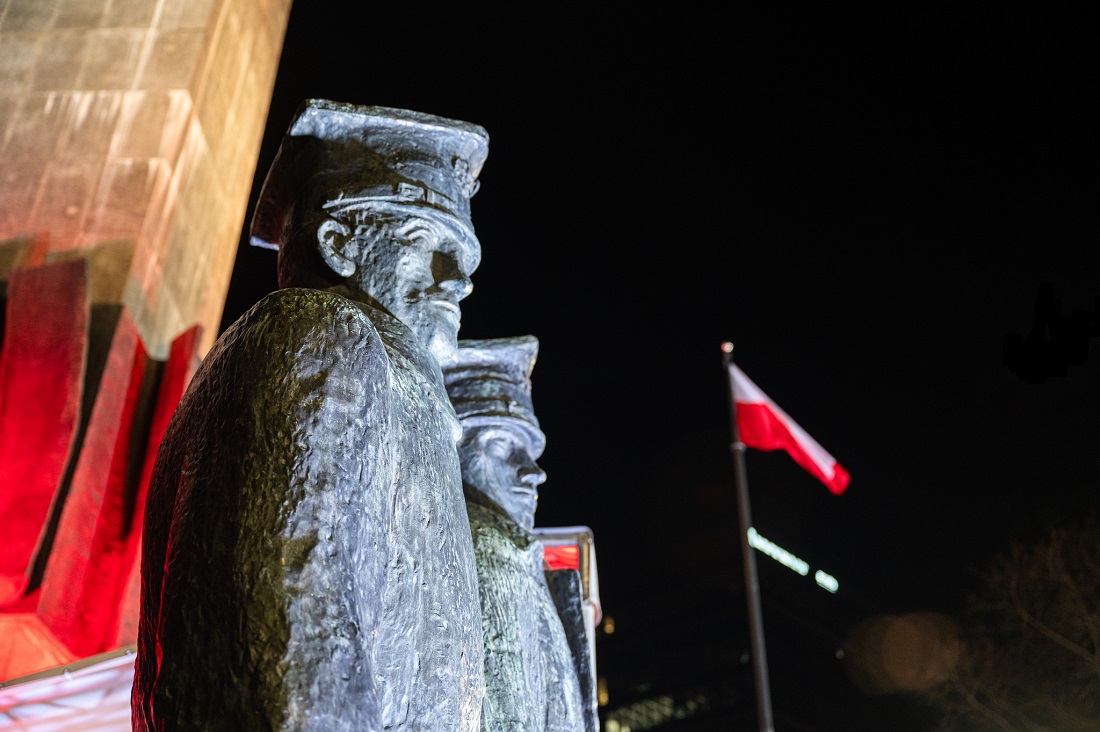 Fragment Pomnika Powstańców Wielkopolskich - figury dwóch powstańców - na tle nocnego nieba, w tle biało czerwona flaga na maszcie - grafika artykułu
