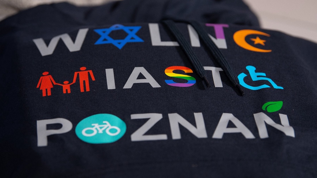 czarna bluza z napisem WOLNE MIASTO POZNAŃ, niektóre litery zapisane w formie znaków graficznych takich jak gwiazda Dawida, półksiężyc, tęczowa flaga, symbol osób niepełnosprawnych, znak rowerowy - grafika artykułu