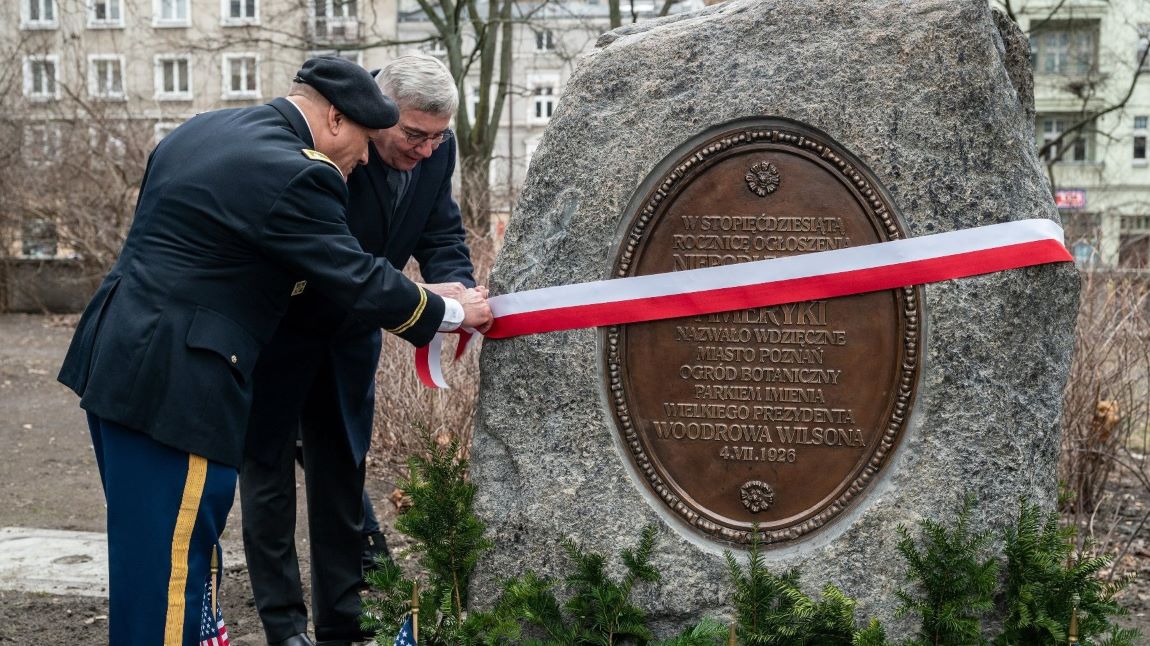 Dwóch męższczyzn, jeden w mundurze armii USA odsłaniają tablicę pamiątkową poświęconą prezydentowi USA Wilsonowi, tablica umocowanajest na głazie, przepasanym biało czerwoną wstęgą - grafika artykułu
