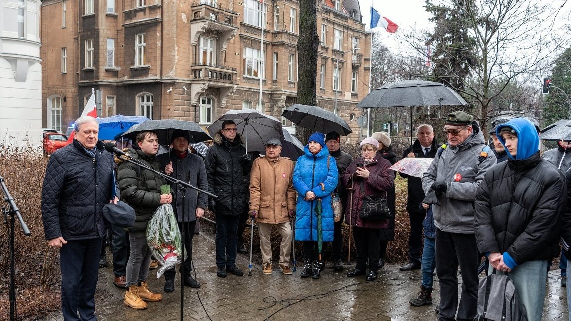 grupa ludzi na skwerze uczestniczących w uroczystości, kobiety i mężczyźni w różnym wieku, trzymają parasowem jest deszczowa pogoda