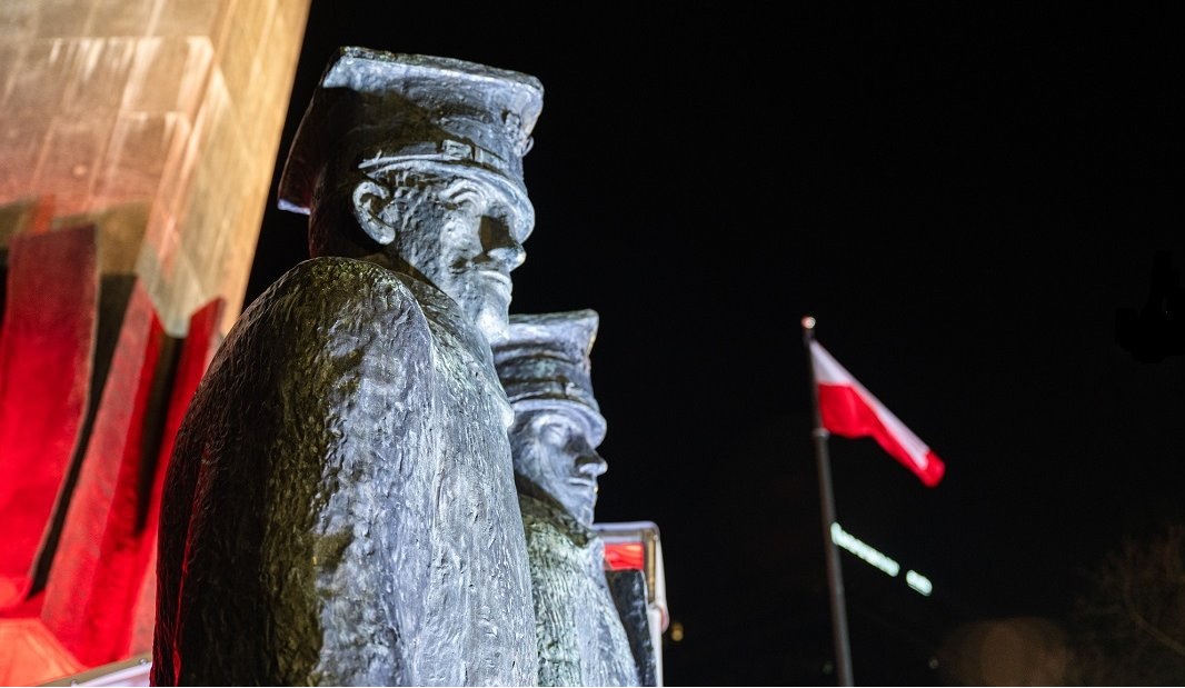 Fragment Pomnika Powstańców Wielkopolskich - figury dwóch powstańców - na tle nocnego nieba, w tle biało czerwona flaga na maszcie - grafika artykułu