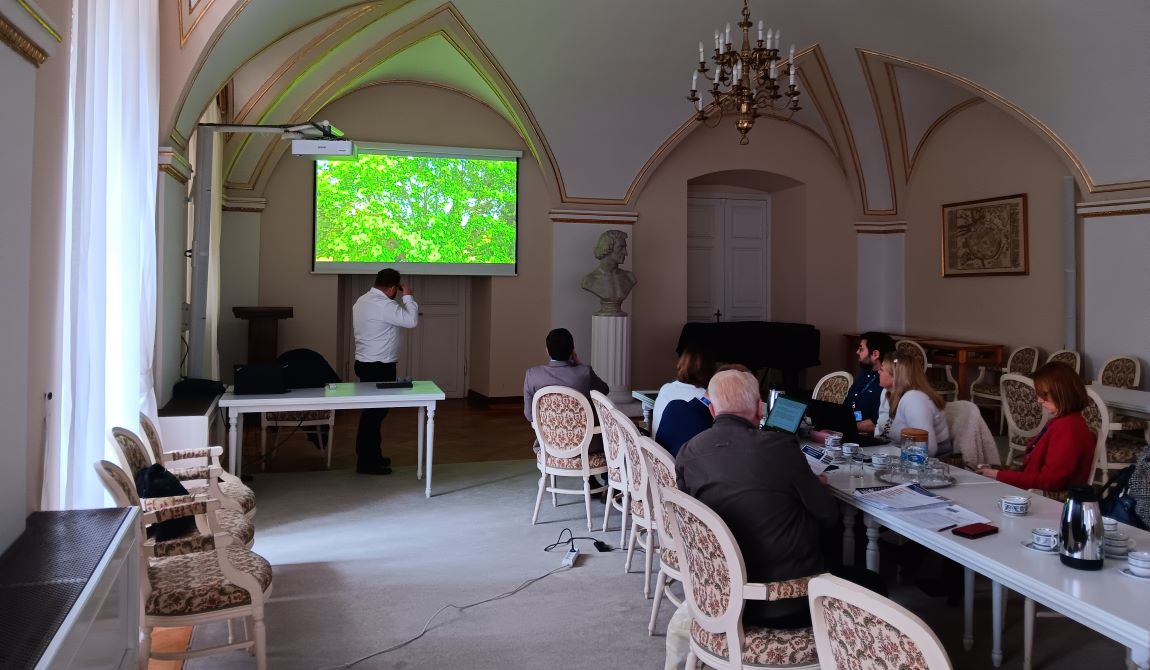 grupa osób kobiet i mężczyzn siedzi przy podłużnyn stole po obu jego stronach, jedna osoba mężczyzna stoi przed ekranem, na którym pokazywana jest prezentacja - grafika artykułu