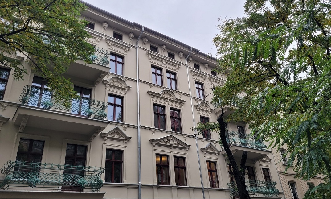 wysoka stara kamienica z odnowioną fasadą, wysokie wąskie okna, balkony ze stalową zdobiona balustradą, przed kamienicą dwa wysokie, rozłożyste drzewa - grafika artykułu