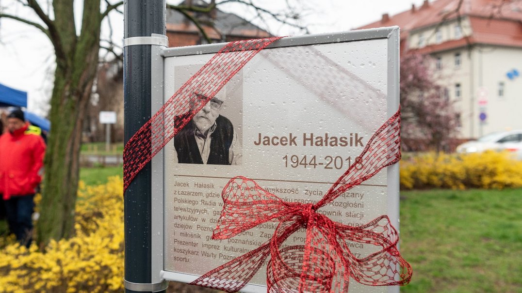 Tablica z wizerunkiem Jacka Hałasika i krótkim biogramem, przytwierdzone do słupka, tablica przewiązana jest czerwoną, ażurową wstążką, za nią skwer z trawnikiem i niską roślinnością, drzewo, ludzkie postaci, dalej kamienice