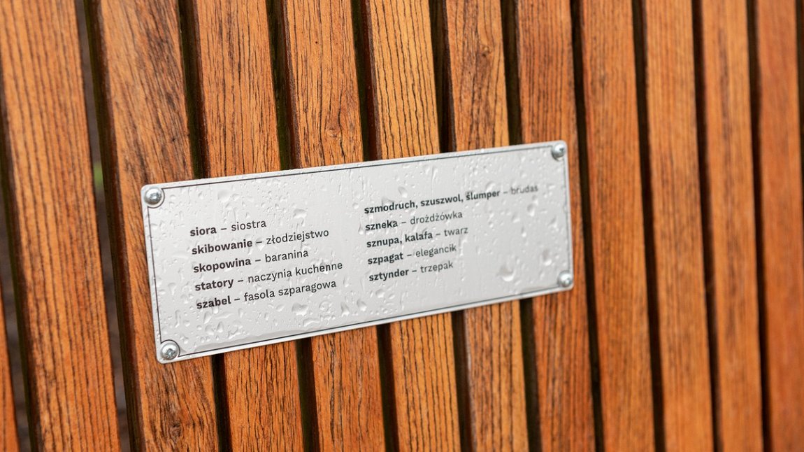 drewniane opracie ławki, a na nim tabliczka z wybranymi słowami w języku polskim i tłumaczeniu na gwarą poznańską