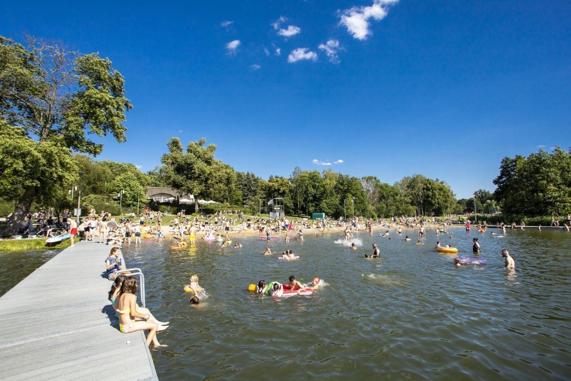 kąpielisko nad jeziorem, plaża, pomost, ludzie siedzą na pomości, kąpią się, leżą na plaży, za plażą drzewa - grafika artykułu