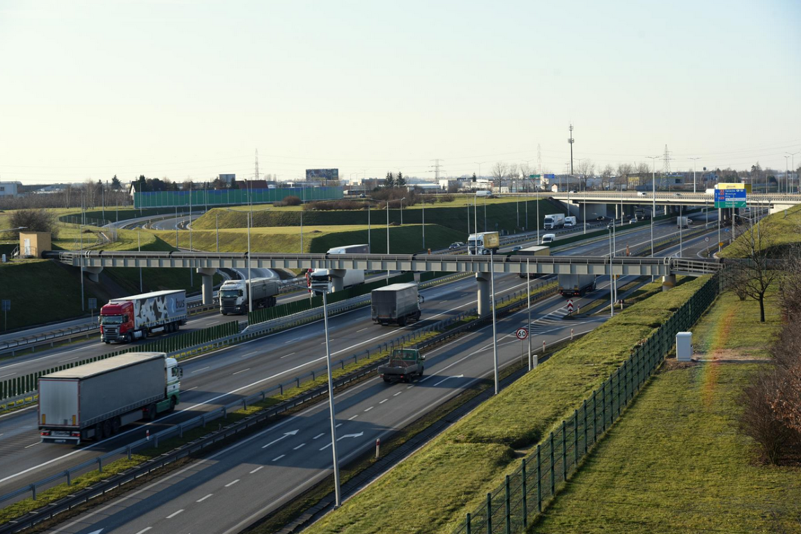 fragment autostrady, pasy ruchu, drogowskazy, pobocze i skarpa porośnięcte trawą, na jezdni samochody osobowe i ciężarowe - grafika artykułu