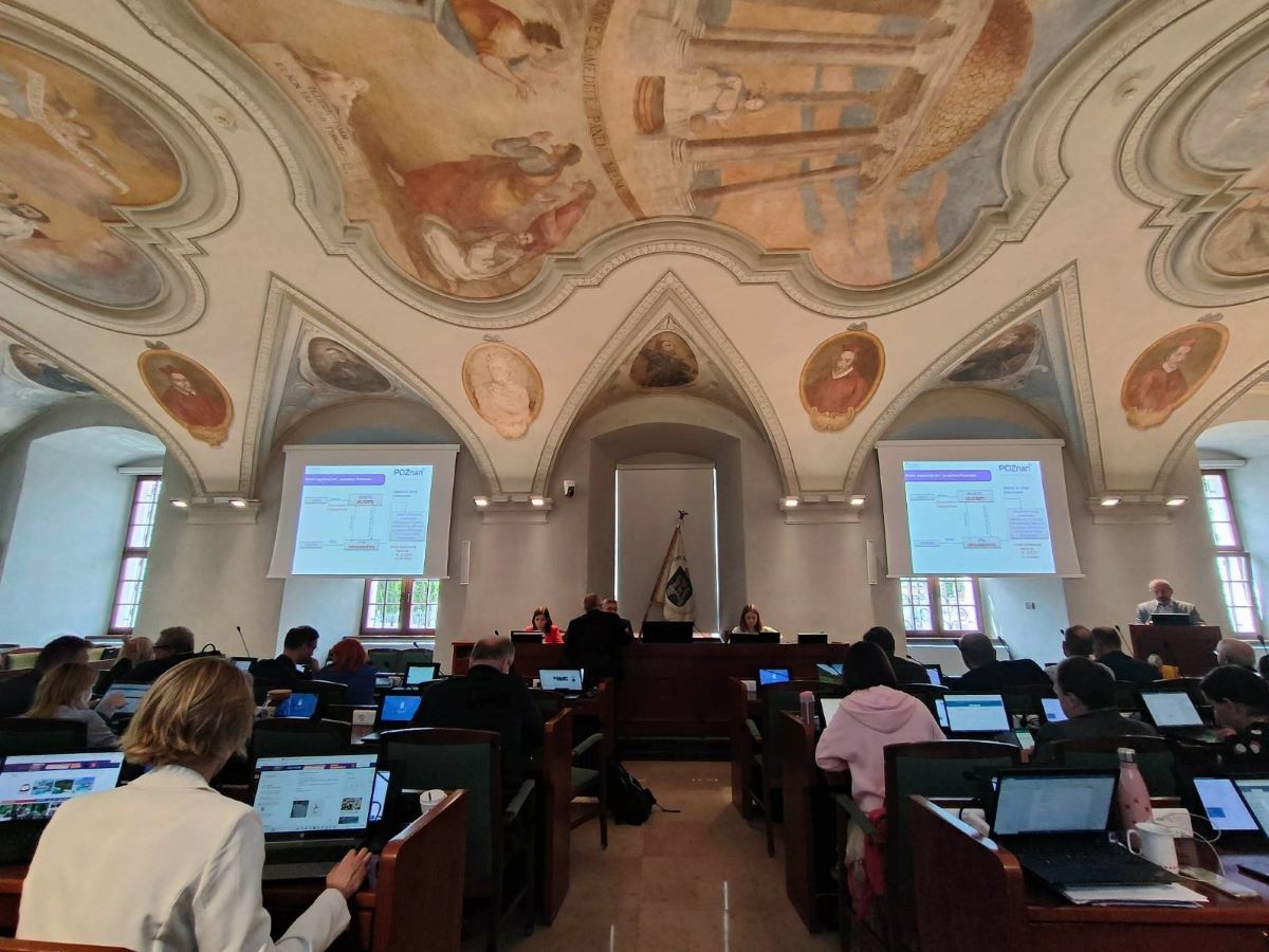 sala z malowanymi sklepieniami, stół prezydialny, za nim siedzą ludzie, na przeciwko stołu rzędy z miejscami dla radnych, stanowiska wyposażone w monitory i mikrofony - grafika artykułu