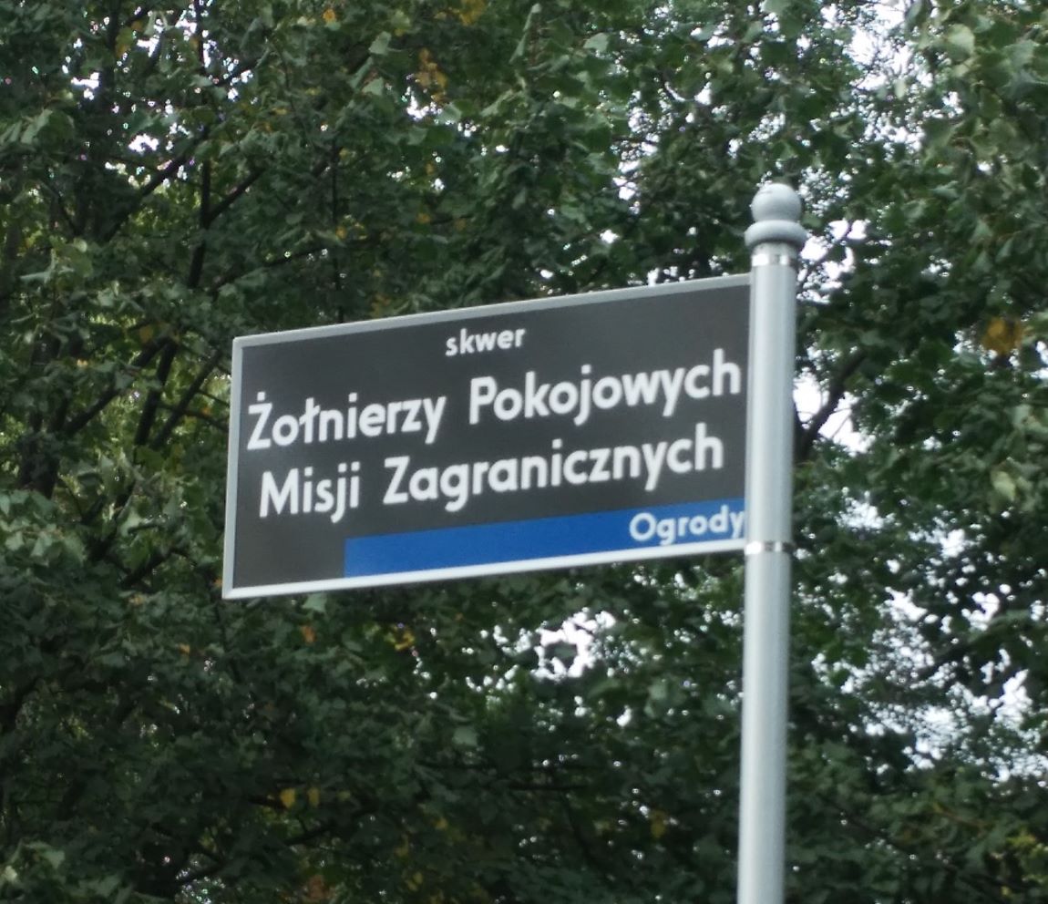 prostokątka tablica z napisem Żołnierzy Pokojowych Misji Zagranicznych umocowane na słupku, za nią zielone korony drzew - grafika artykułu