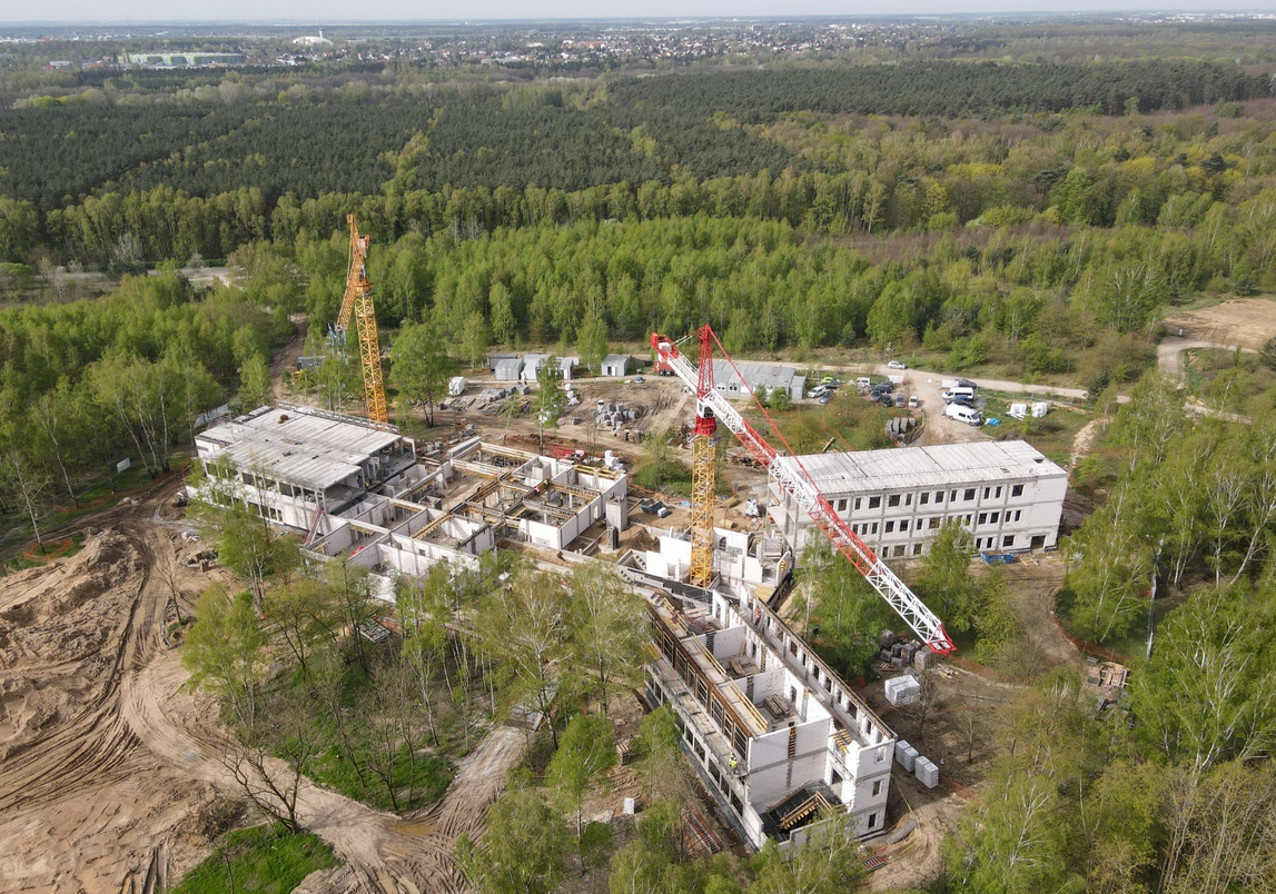 teren budowy z lotu ptaka, budynki w stanoe surowym, dżwigi budowlane, fragmenty, wokół gęsto rosnące drzewa - grafika artykułu