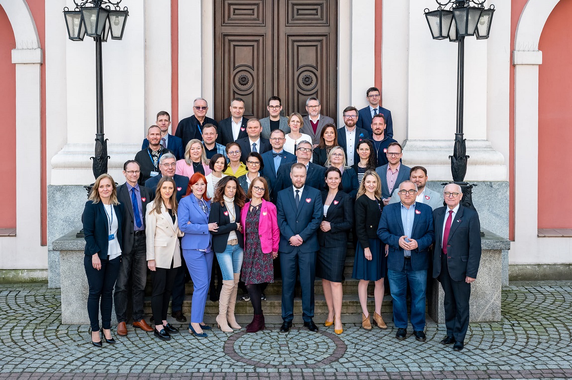 grupa kilkudziesięciu osób kobiet i mężczyzn w różnym wieku ustawiona w kilku rzędach na schodach przed budynkiem - grafika artykułu