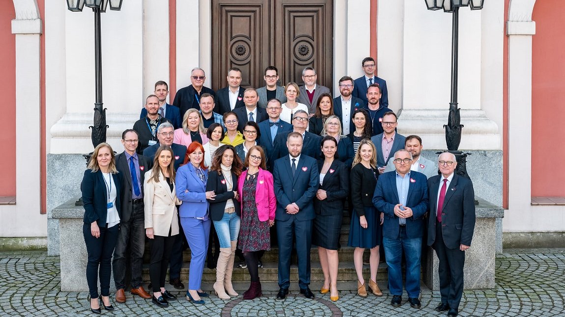 grupa kilkudziesięciu osób kobiet i mężczyzn w różnym wieku ustawiona w kilku rzędach na schodach przed budynkiem